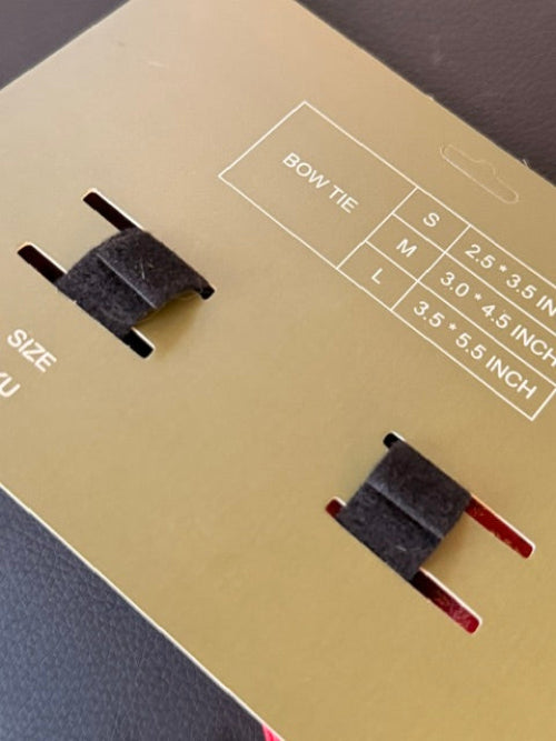 A close-up of a gold bow tie display card showing two black bow ties, size labels, and printed measurements for sizes S, M, and L.