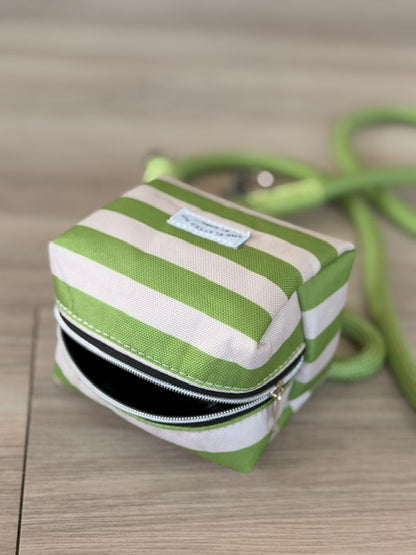 A small zippered pouch with green and white stripes, featuring a light blue label and a matching green strap, resting on a light wood floor.