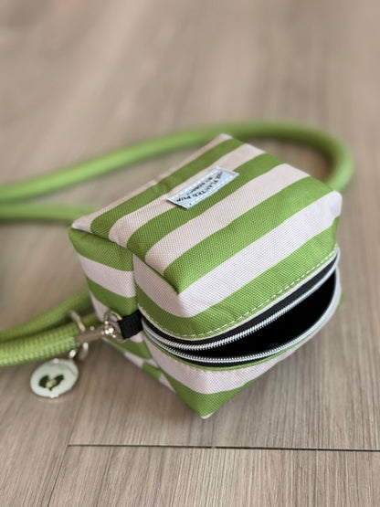 A small square zippered pouch with green and white stripes, featuring a matching green strap and silver zipper, resting on a light wood floor.