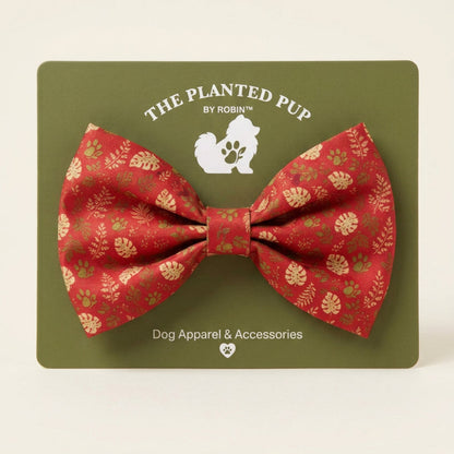 A red bow tie with a gold leaf and paw print pattern, displayed on its olive green backing card from 'The Planted Pup by Robin' on a speckled granite countertop.