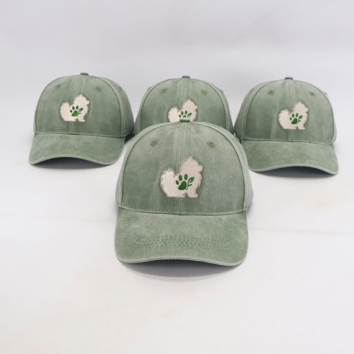 Four sage green baseball caps arranged on a white background, each featuring an embroidered white silhouette of a small dog with a green paw print on the front.