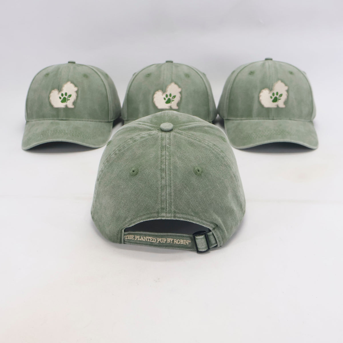 Three sage green baseball caps with a white and green fluffy dog patch and paw print on the front, displayed on a white background with one cap shown from the back.