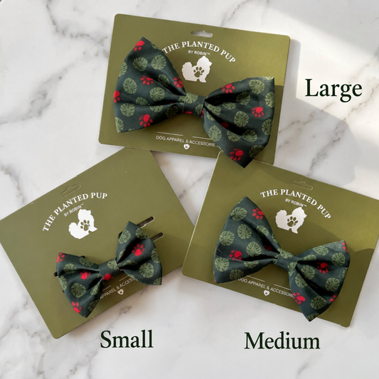 Three dog bow ties from The Planted Pup by Nature's Store shown on olive green retail cards, labeled Small, Medium, and Large, arranged on a white marble surface. The bow ties have a dark green base with red paw prints and light green leaf patterns.