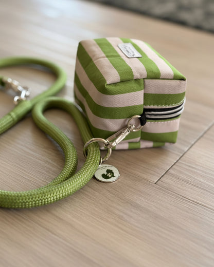 A small square zippered pouch with green and cream horizontal stripes, featuring a matching green rope handle and a silver metal clasp with a round hanging tag, shown on a light wood floor.